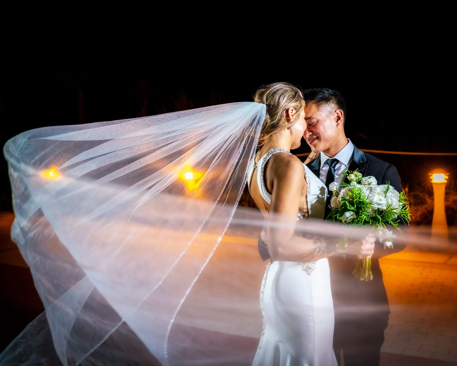 Bride and groom beneath a wind-blown veil at night